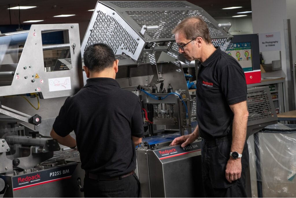 Two men collaborating on a machine in a factory setting, focused on their task and surrounded by industrial equipment.