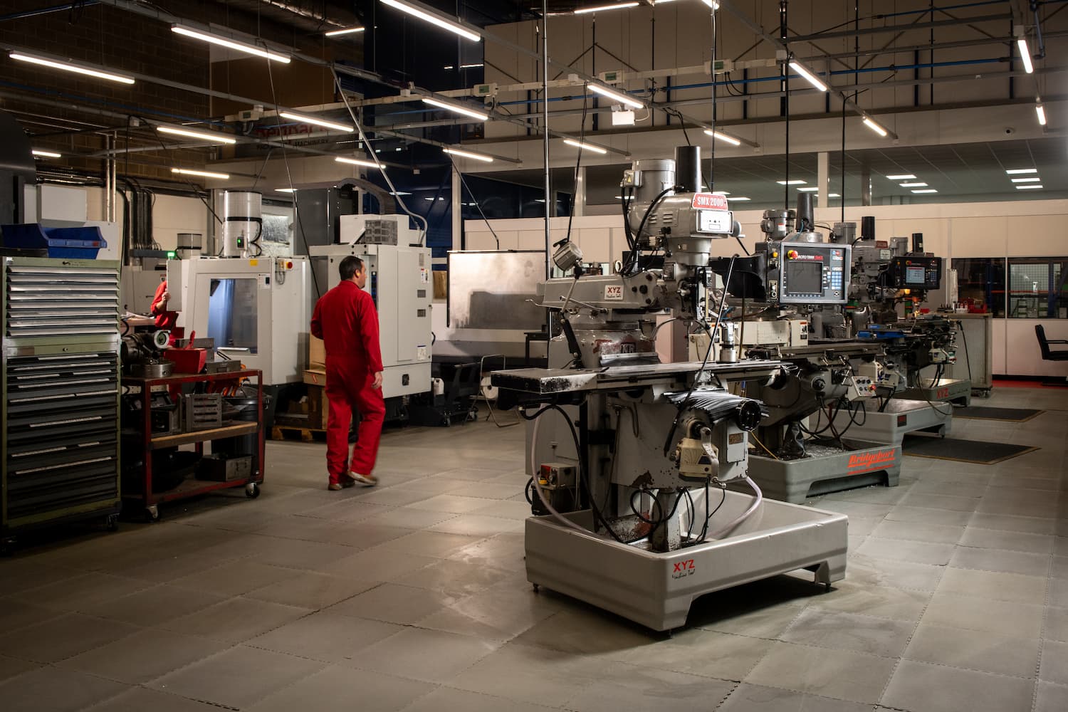 In a machine shop filled with equipment, a man wearing a red uniform stands, focused on his work environment.